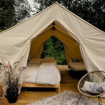 A woman in a nude bathingsuit walks toward a large white bell tent in a grassy backyard, with a small wooden sauna on a trailer to the right and trees in the background.