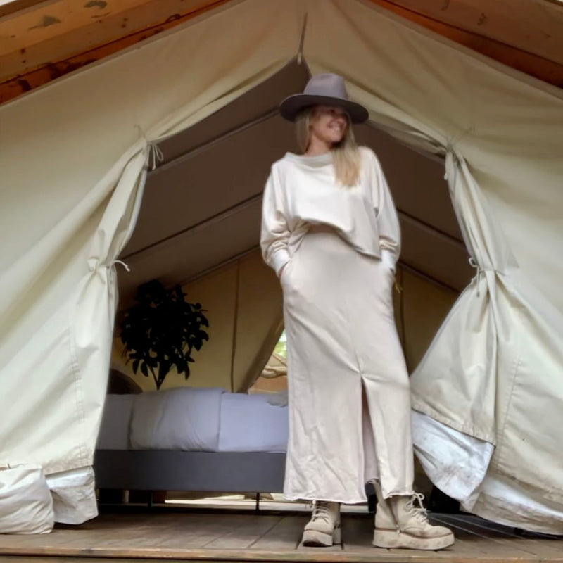 Person wearing a cream sweatshirt + Skirt standing in front of a glamping tent with trees in the background