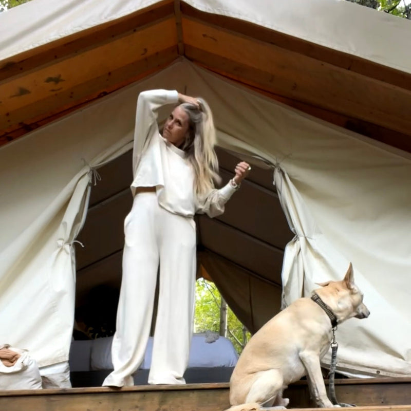 Woman in cream outfit standing in front of a safari tent with a dog sitting on the ground.