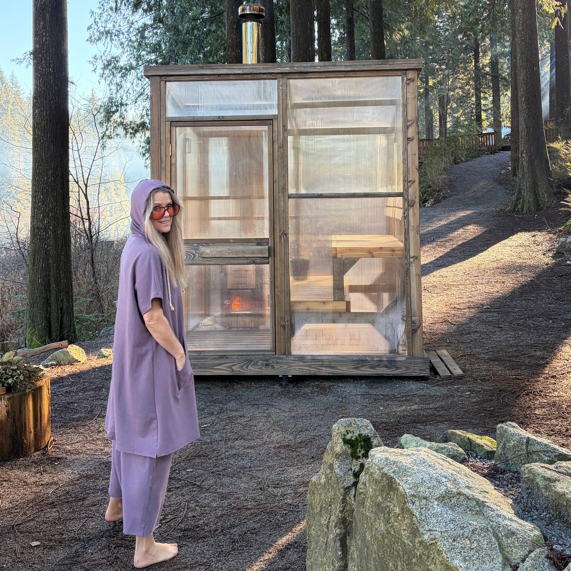 Person in a purple outfit standing next to a small wooden sauna in a forest setting.