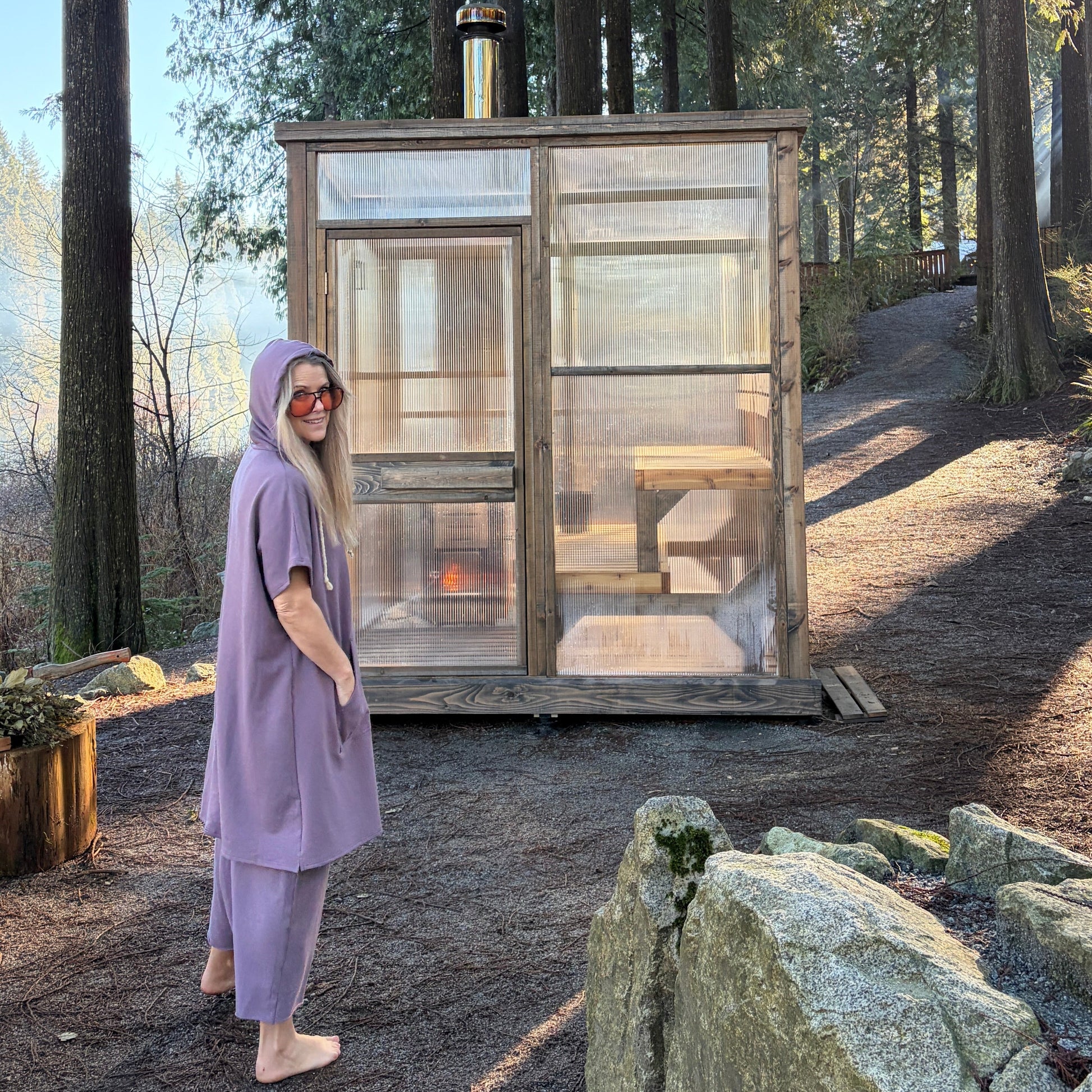 Person in a purple outfit standing next to a small wooden cabin sauna in a forest setting.