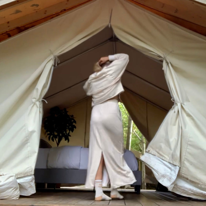 Person in a cream outfit standing in a large glamping tent with a bed inside, surrounded by trees.