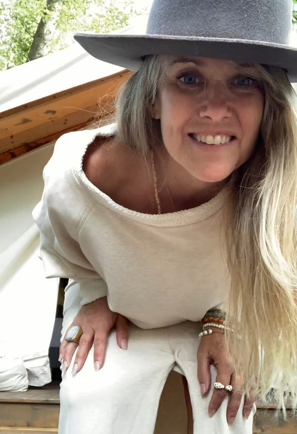 Woman wearing a gray hat and cream sweatshirt slightly off the shoulder outfit with a blurred glamping background