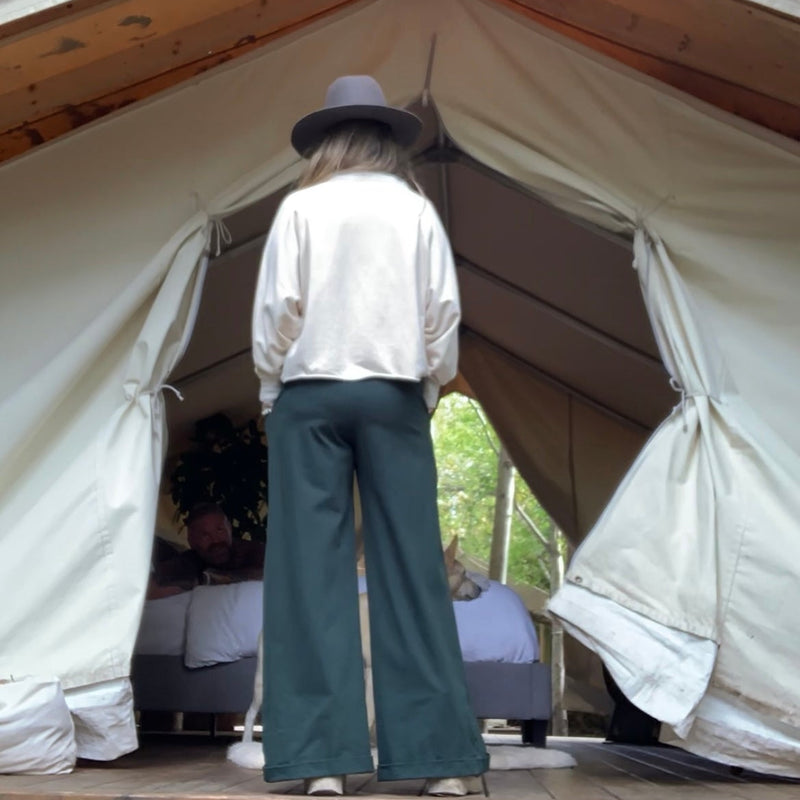 Person standing at the entrance of a large canvas tent with a wooden deck, surrounded by trees, wearing a Cream Sweatshirt + Green Trousers