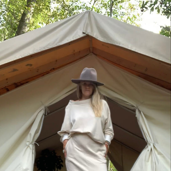 Woman in a cream outfit [ sweatshirt + sweat skirt ] and hat standing in front of a large tent with trees in the background