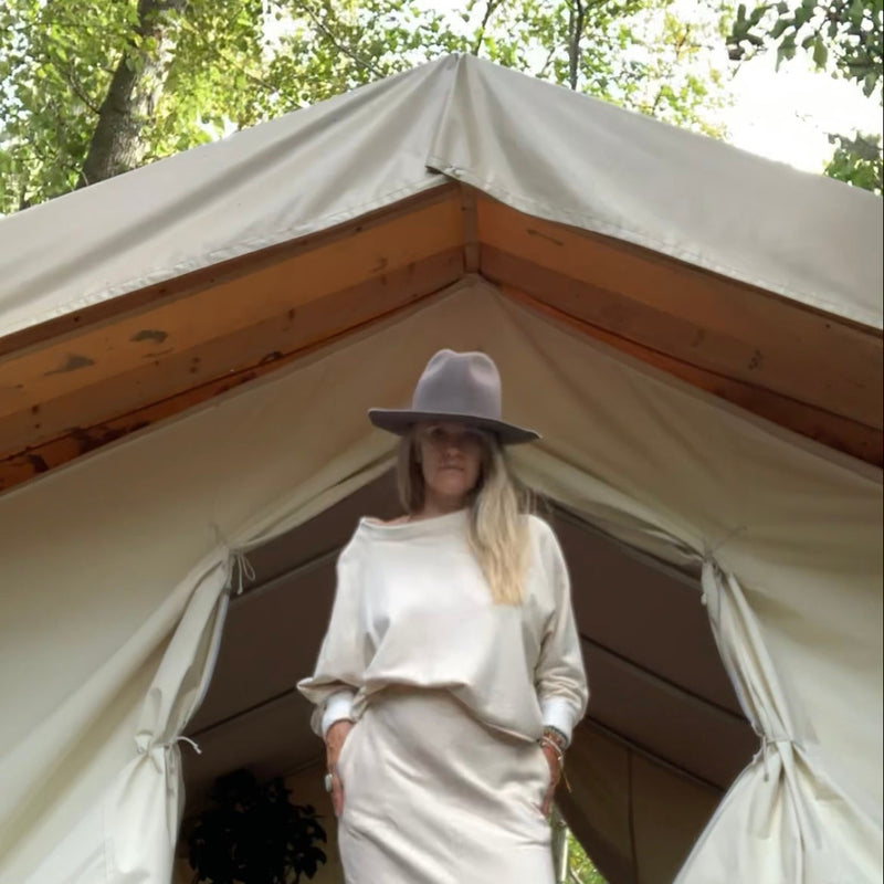 Woman in a cream outfit [ sweatshirt + sweat skirt ] and hat standing in front of a large tent with trees in the background