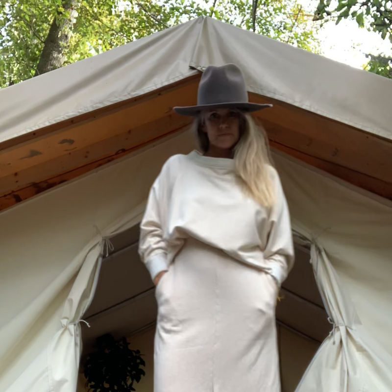 Woman wearing a cream outfit standing in front of a large glamping tent with wood structure & trees in the background