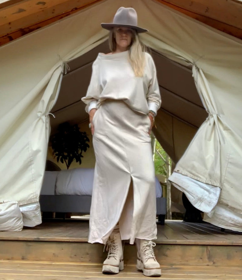 Person wearing a hat, in a cream outfit standing in front of a large safari tent with a wooden floor & bed & plant inside