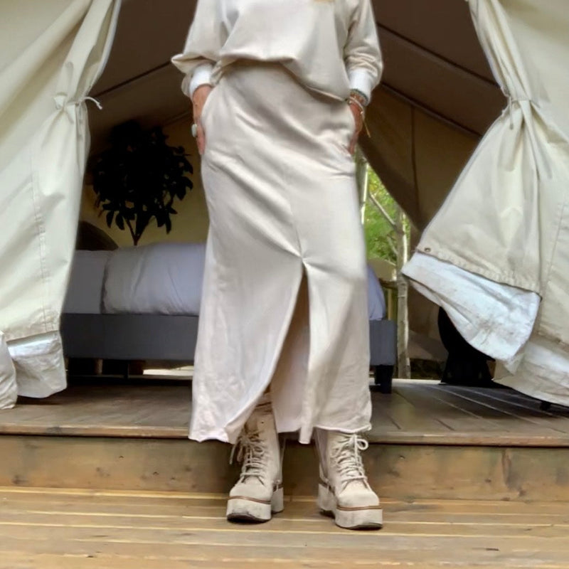 Person in a cream outfit standing in front of a large safari tent with a wooden floor & bed & plant inside
