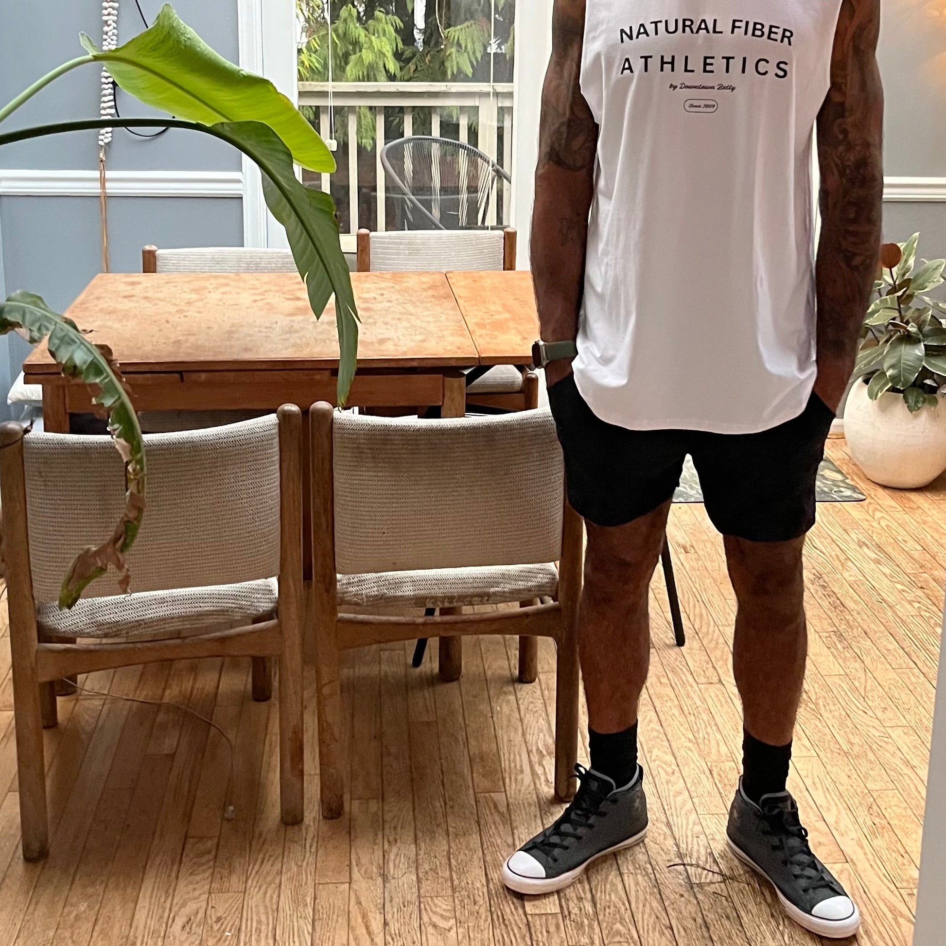 A man wearing a white tank top with the text 'Natural Fiber Athletics' and a pair of black gym shorts standing in a room with wooden flooring and a plant in the background.