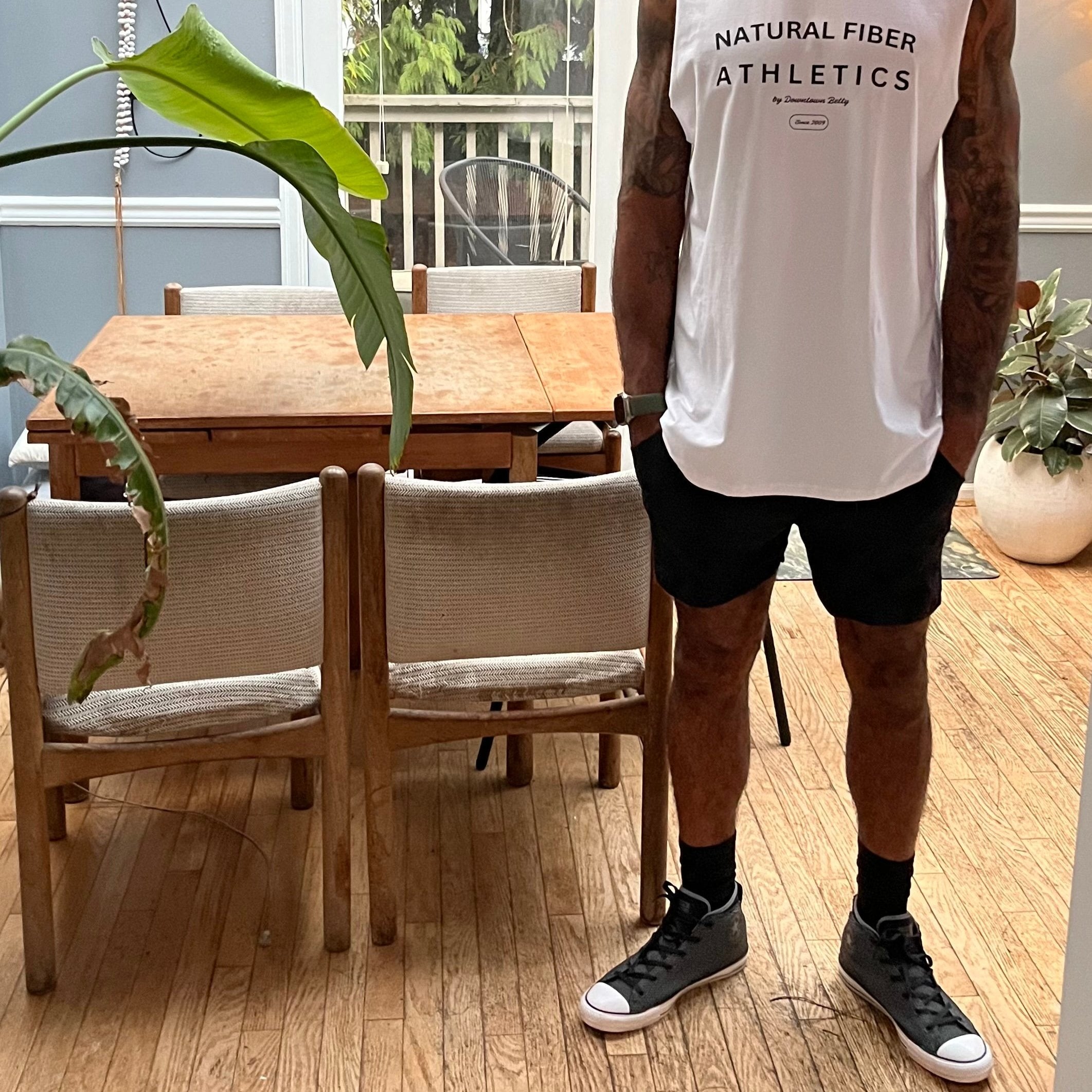 A man wearing a white tank top with the text 'Natural Fiber Athletics' and a pair of black gym shorts standing in a room with wooden flooring and a plant in the background.