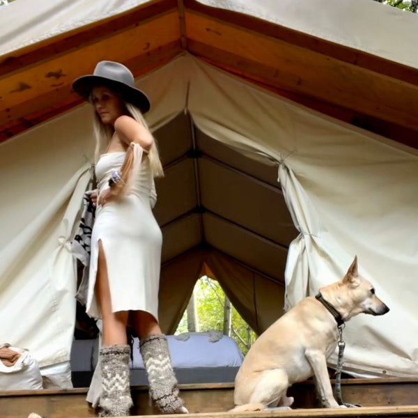 Woman in a cream dress and hat standing next to a dog in front of a large canvas glamping tent.