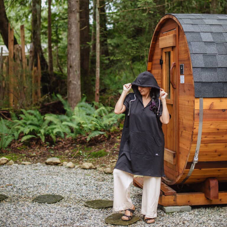 Person wearing a Black Poncho hoody standing in front of a wooden barrel-style cabin in a forest setting