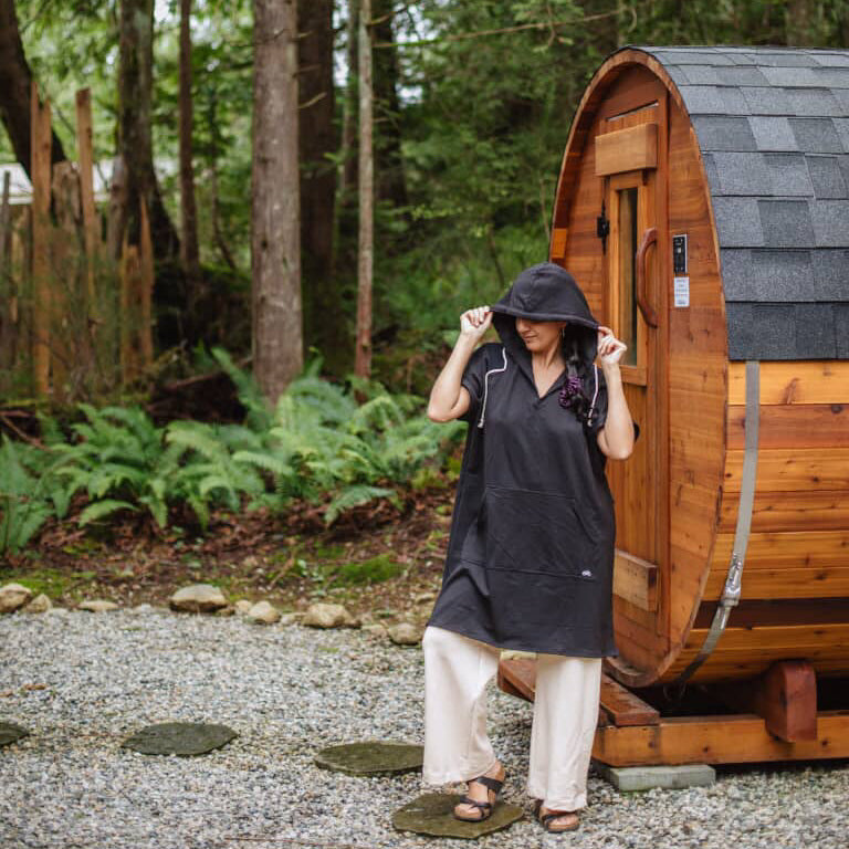 Person wearing a Black Poncho Hoody & Cream Pants, standing in front of a wooden barrel-style cabin in a forest setting