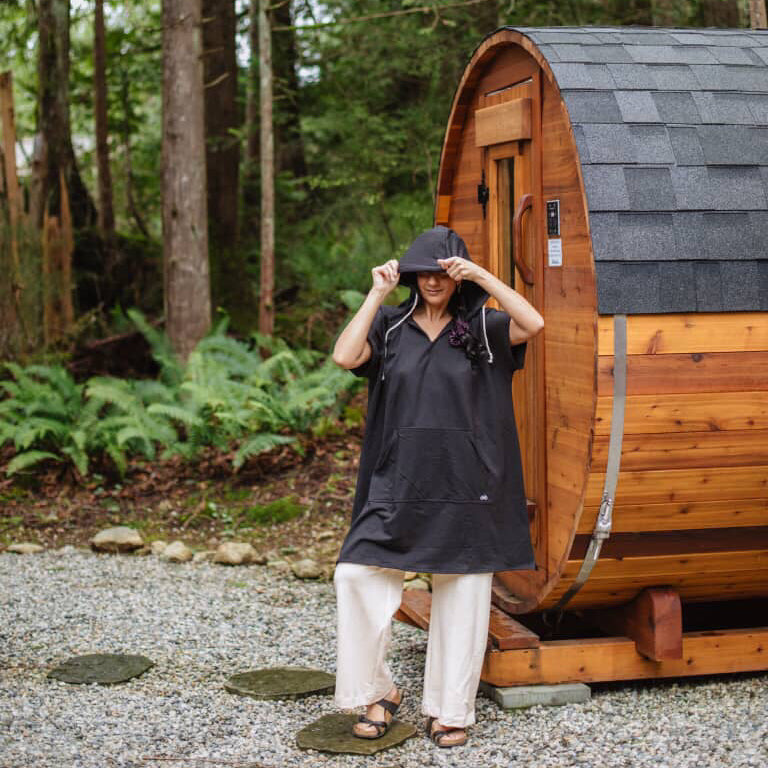 Person standing in front of a wooden barrel-style sauna wearing a Black Poncho Hoody in a forest setting