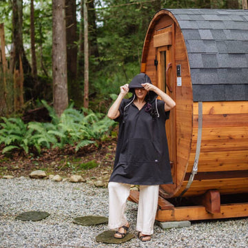 Person standing in front of a wooden barrel-style sauna wearing a Black Poncho Hoody in a forest setting