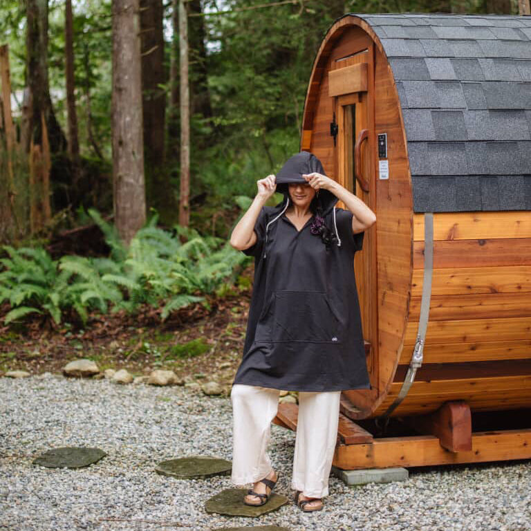 Person wearing a Black Poncho Hoody & Cream Pants, standing in front of a wooden barrel-style cabin in a forest setting