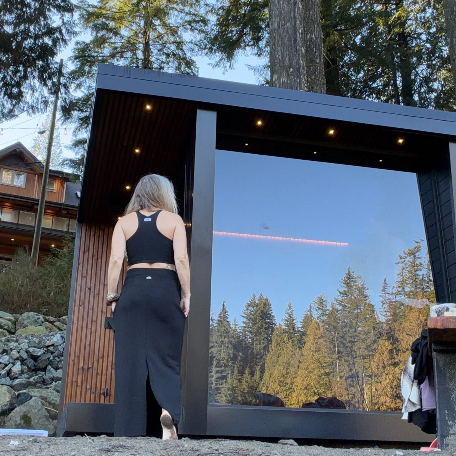 Person standing in front of a modern sauna structure with log cabin & trees in the background