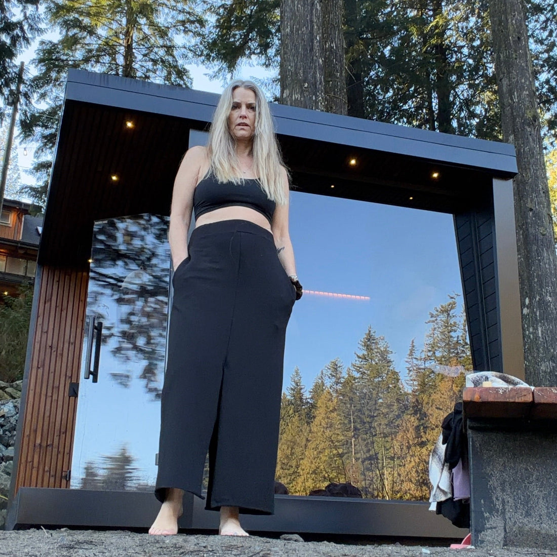 Woman  wearing a black skirt & matching tank top in front of a moder black & wood mirrored sauna with a scenic forest background