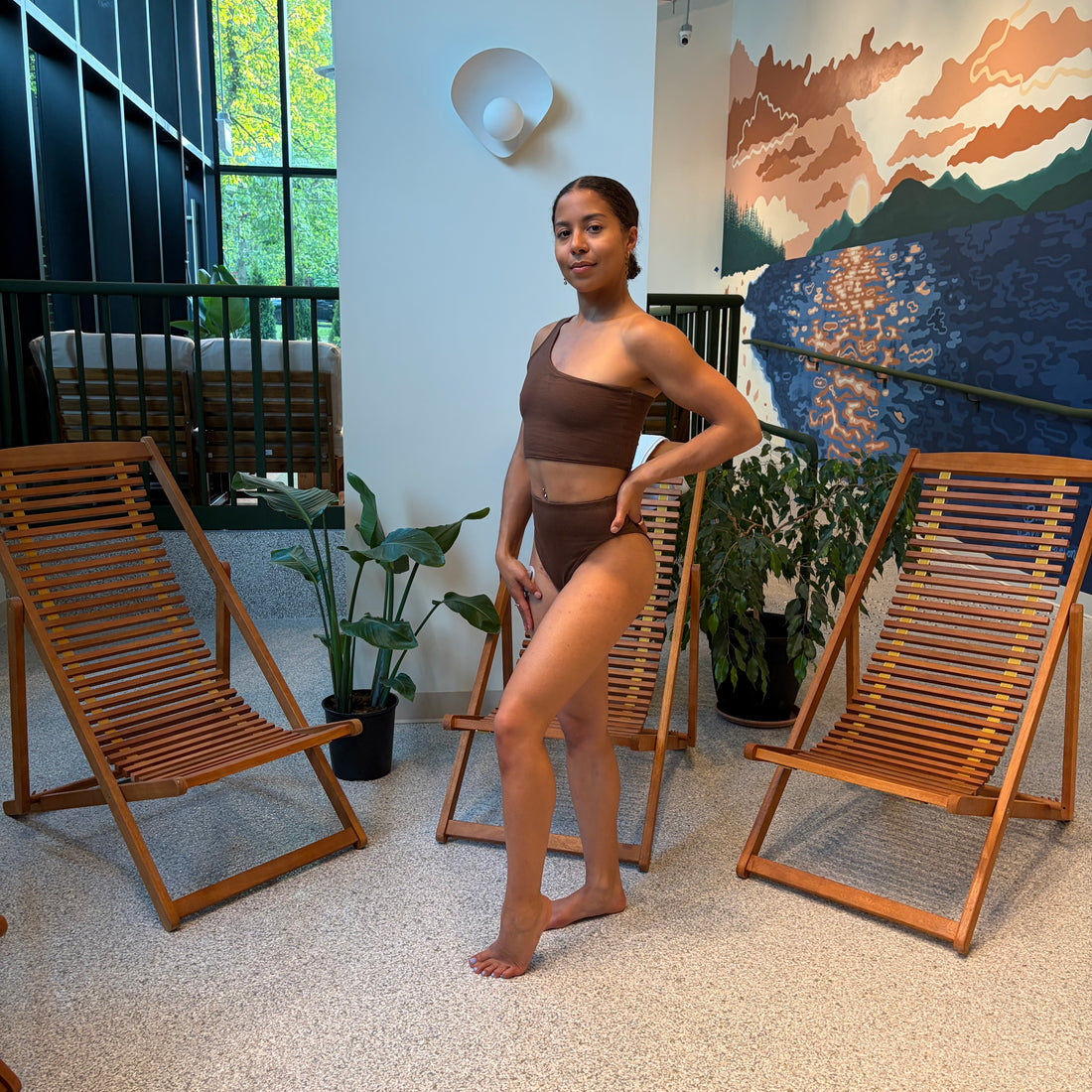 Woman in a brown swimsuit standing in a room with wooden chairs and plants.