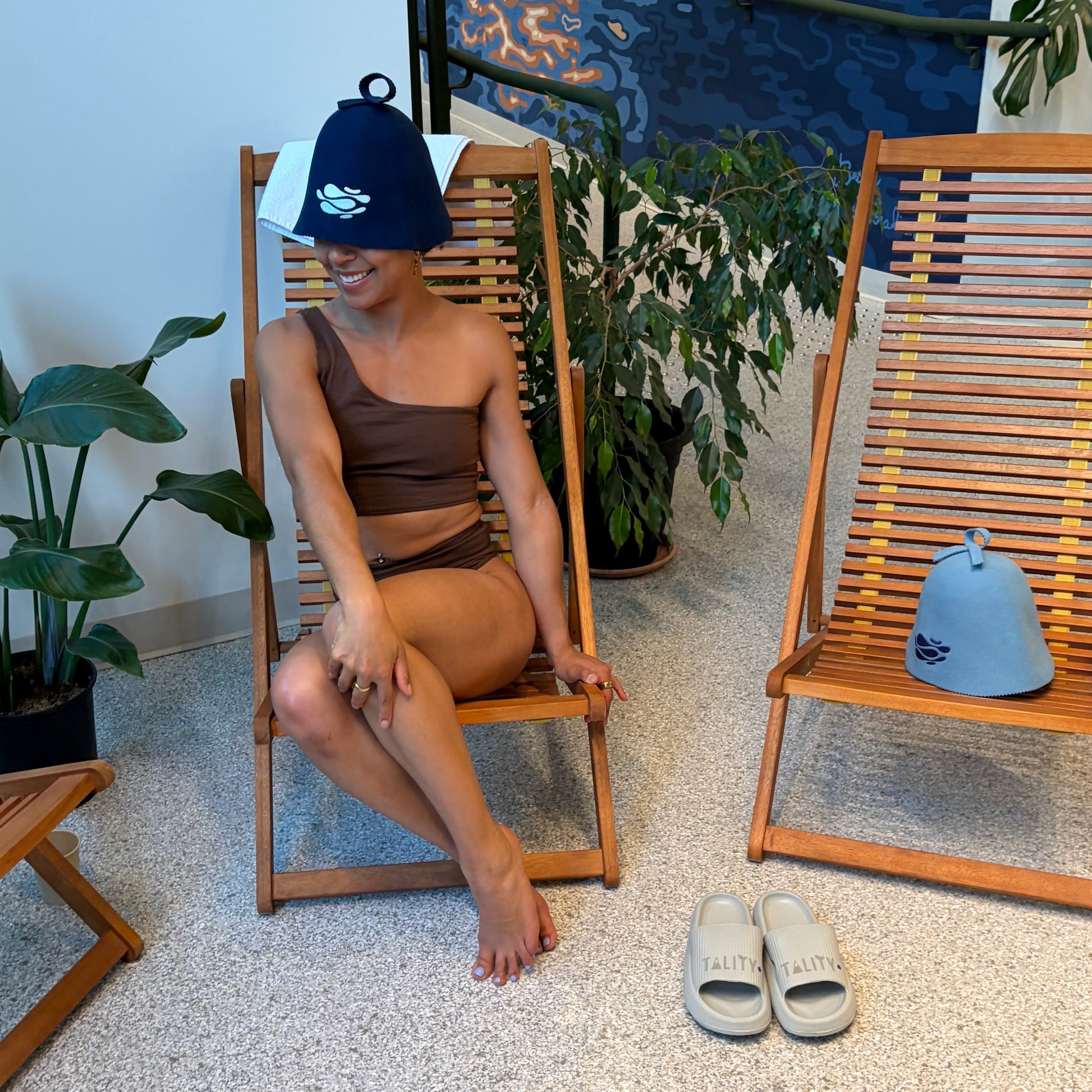 Person wearing a brown bikini, sitting on a wooden chair in a room with plants  a blue wall.