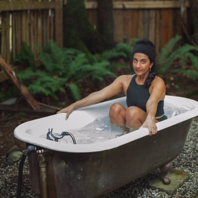 Person sitting in a cold plunge bathtub outdoors wearing a natural fiber Sauna Suit with a wooden fence and greenery in the background