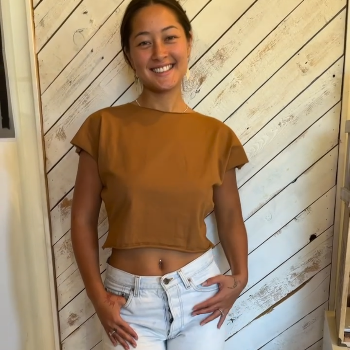 Person wearing a mustard top and white jeans standing against a wooden paneled wall.