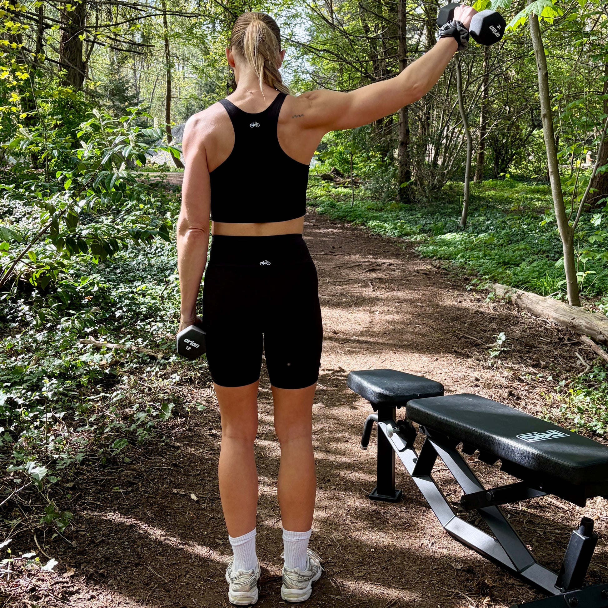 Person in athletic wear standing on a forest path with a bench and camera equipment.