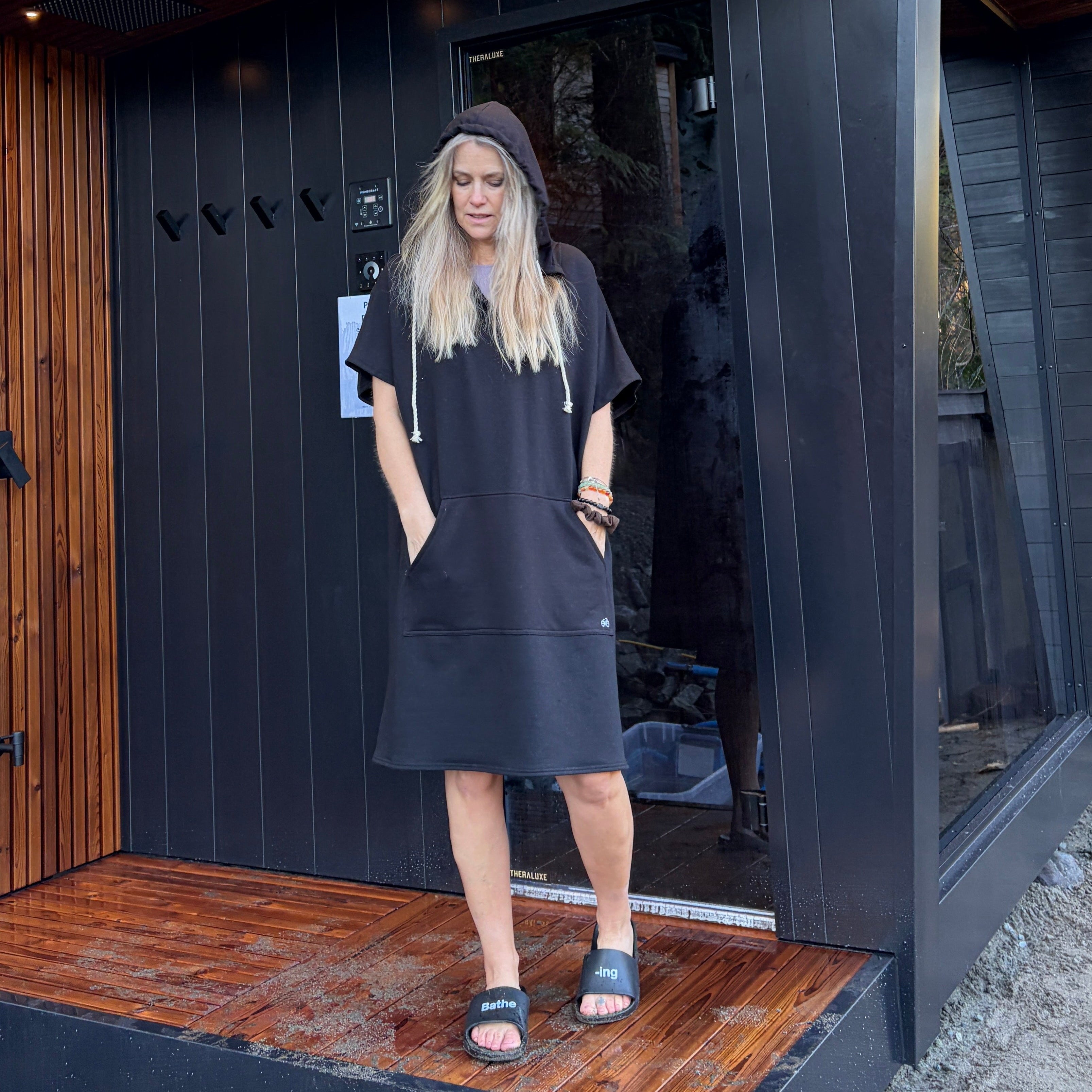 Woman standing in front of a modern black sauna cabin with wooden deck wearing a black Poncho Hoody.