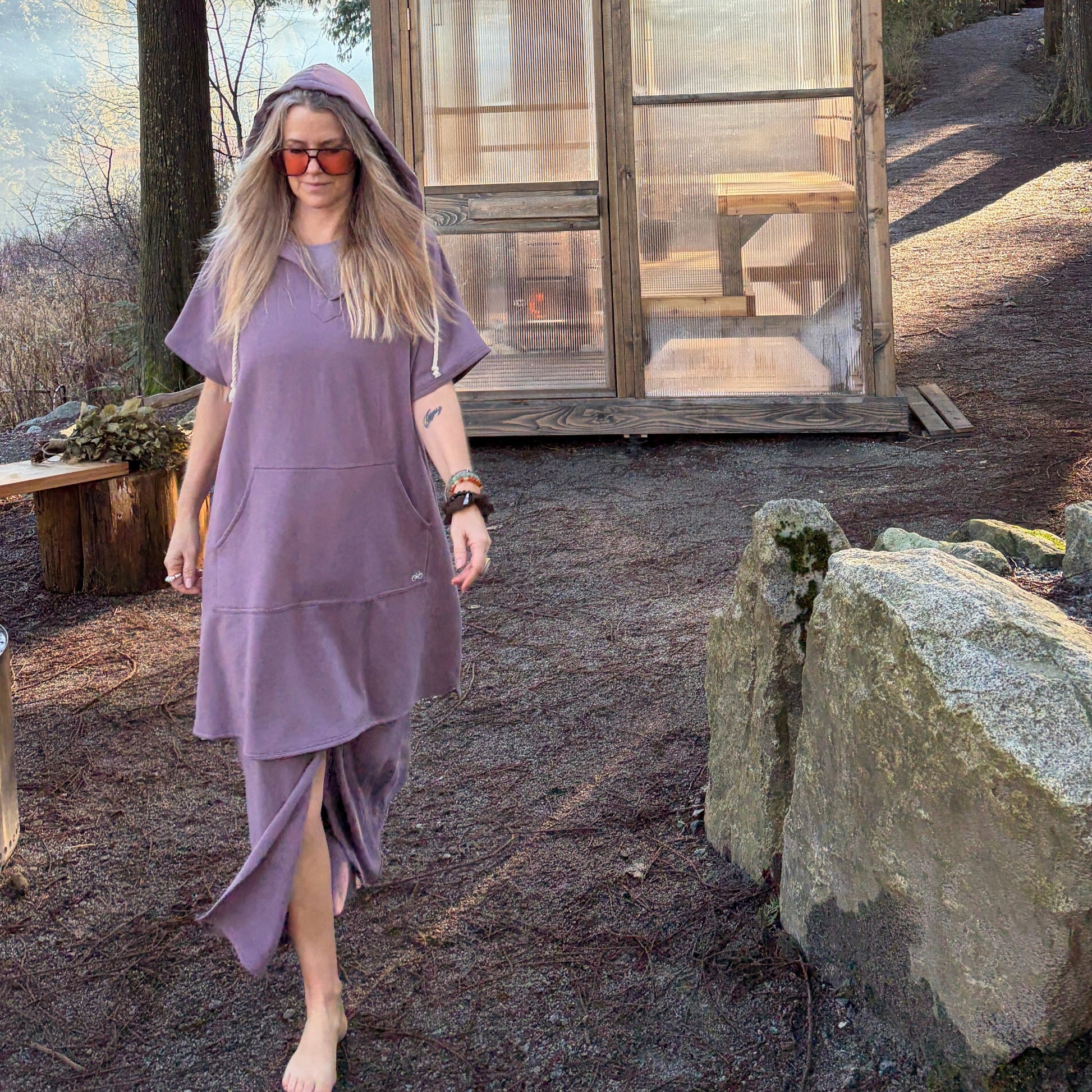 Woman in a purple Poncho Hoody worn over a matching Skirt standing in front of a small wooden see-thru sauna in a forest.