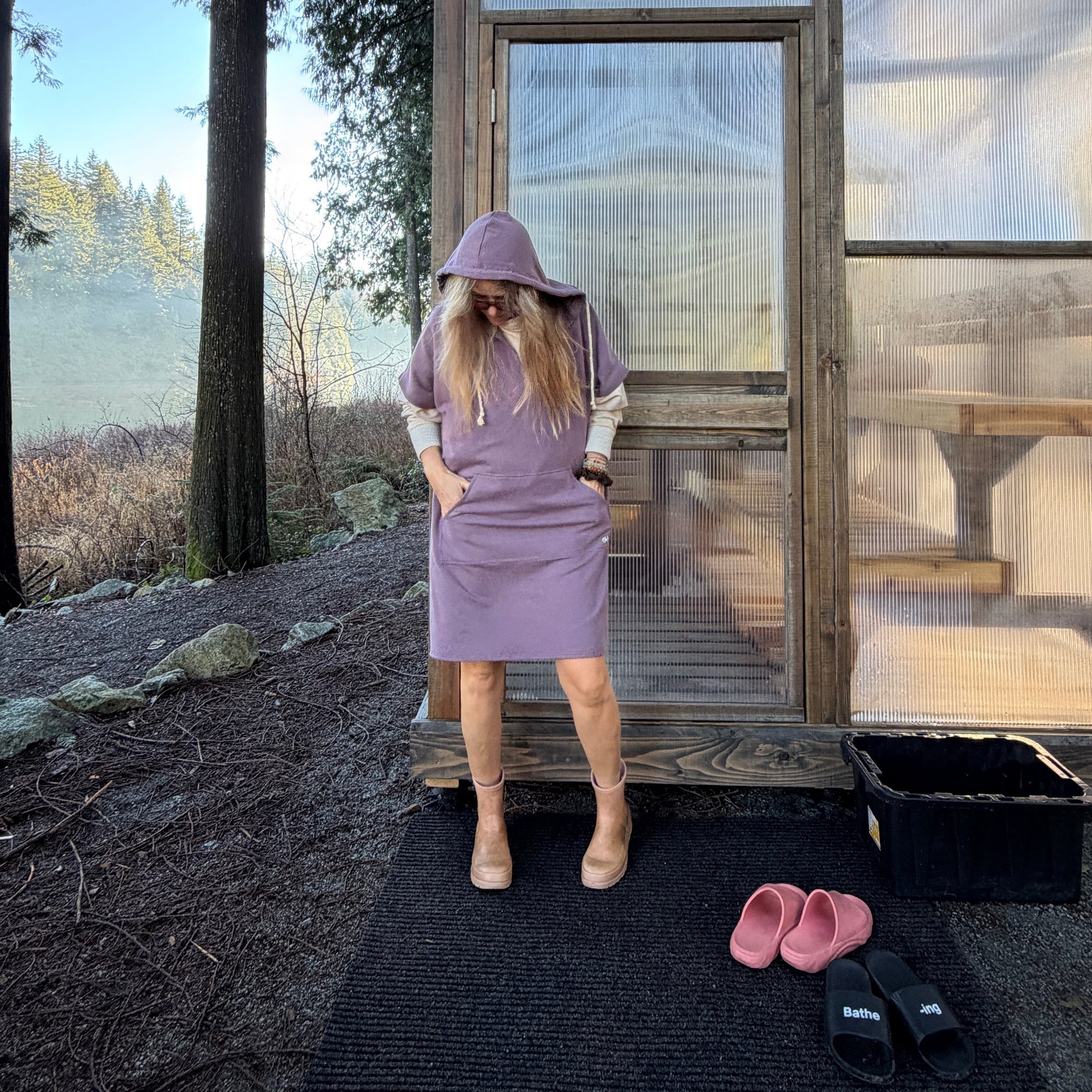 Person in a purple poncho hoody standing in front of a glass sauna cabin with trees in the background