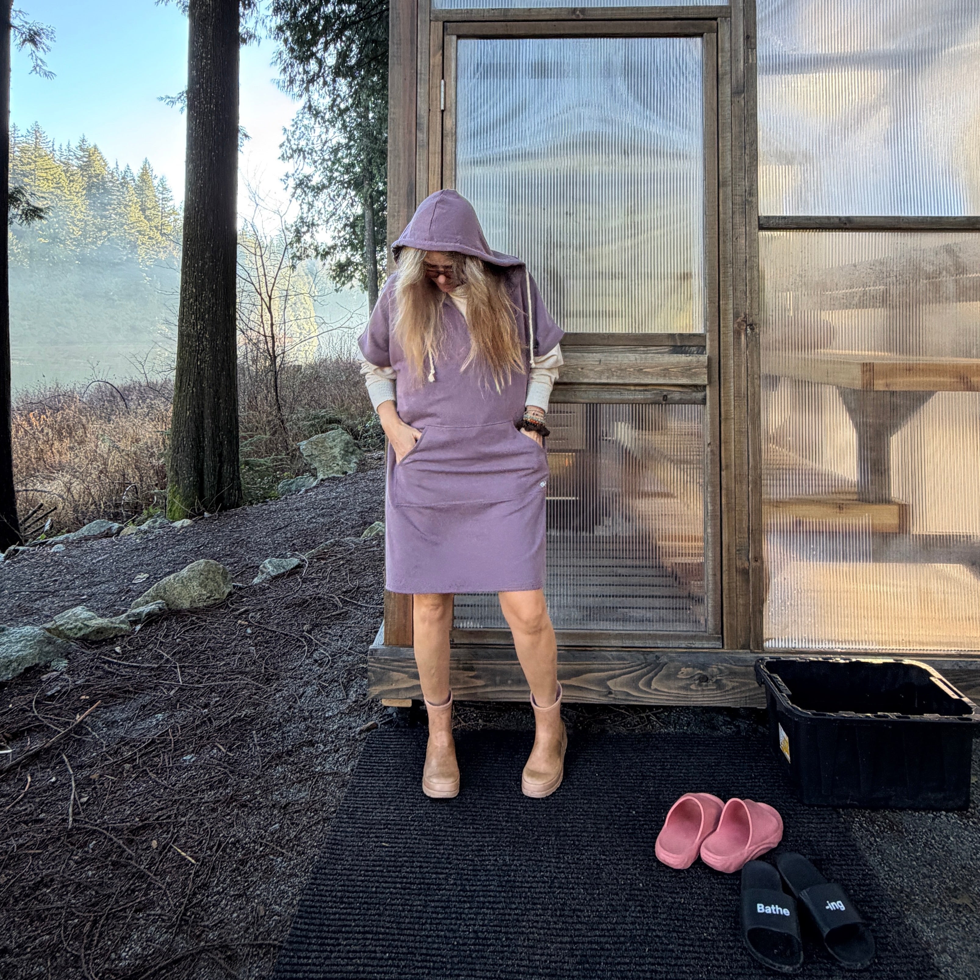 Person in a purple poncho hoody standing in front of a glass sauna cabin with trees in the background