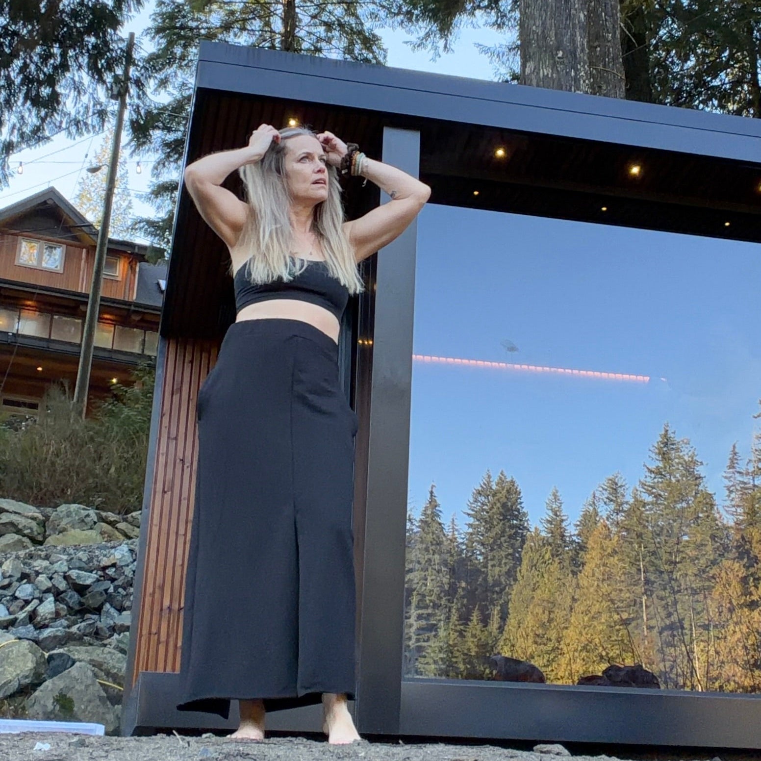 Woman  wearing a black skirt & matching tank top in front of a moder black & wood mirrored sauna with a scenic forest background