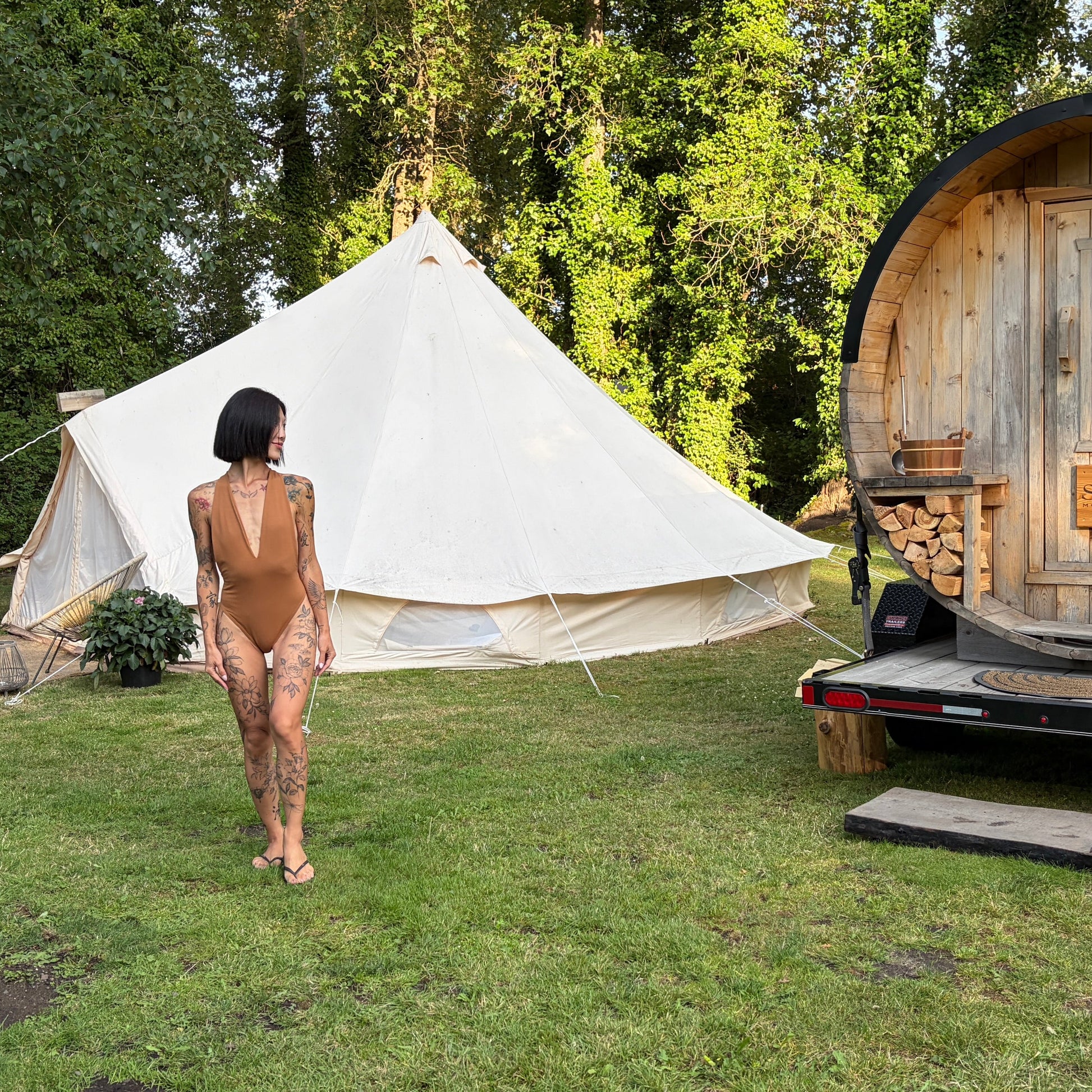 A woman in a nude bathingsuit walks toward a large white bell tent in a grassy backyard, with a small wooden sauna on a trailer to the right and trees in the background.
