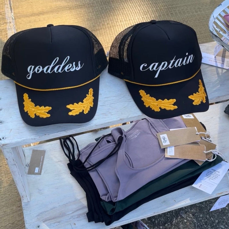 Two black baseball caps with embroidered oak leaves & gold rope, with 'goddess' I 'captain' text side by side on a white shelf @ the Country Club Festival
