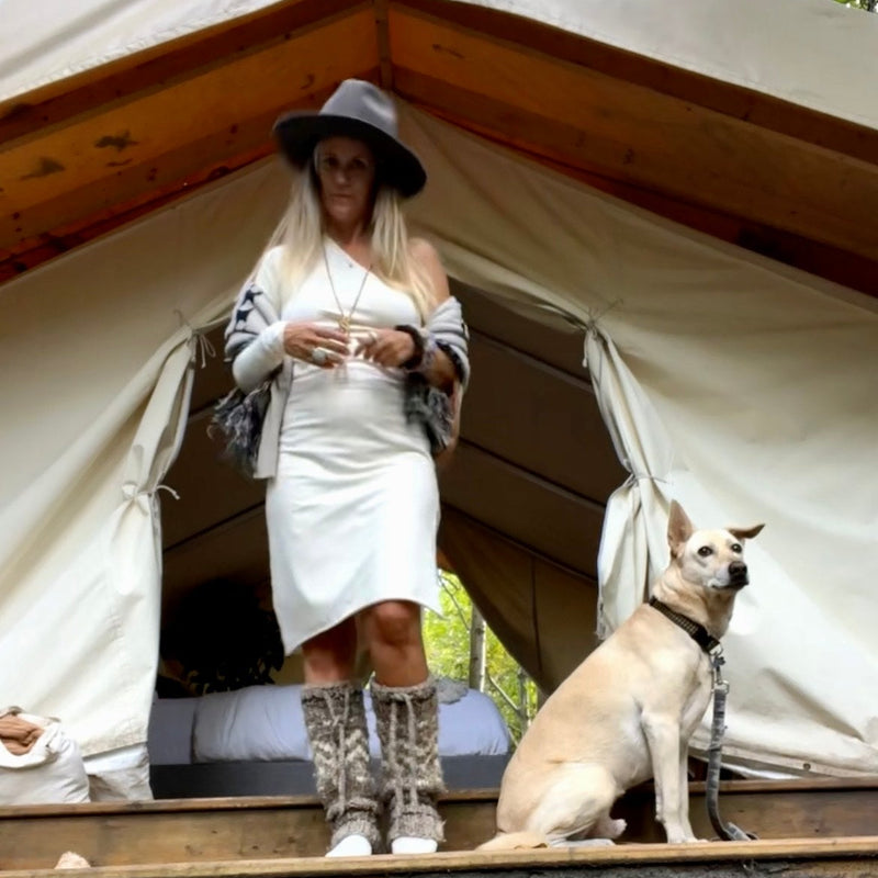 Woman in a cream dress , shawl & a hat standing next to a dog in front of a large tent.