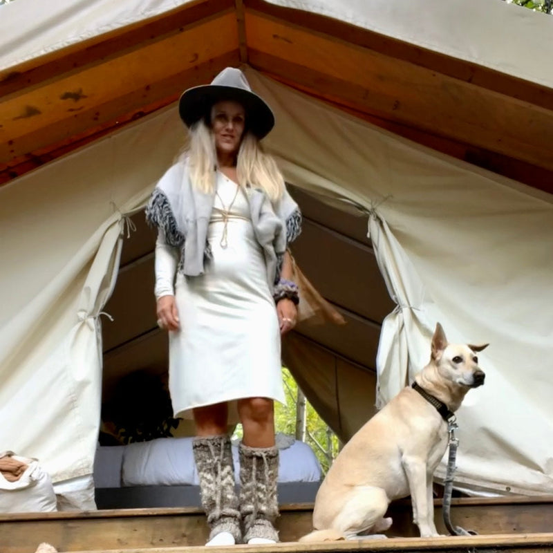 Woman in a cream dress and hat standing in front of a canvas glamping tent with a dog sitting beside her.