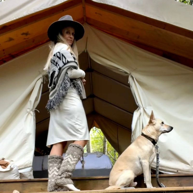 Woman in a hat, poncho wearing  a cream dress standing next to a dog in front of a large tent outdoors
