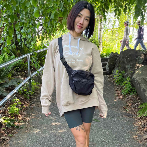 Person wearing a beige hoodie, bike shorts shorts, and white sneakers with a black bag, standing on a path with greenery.