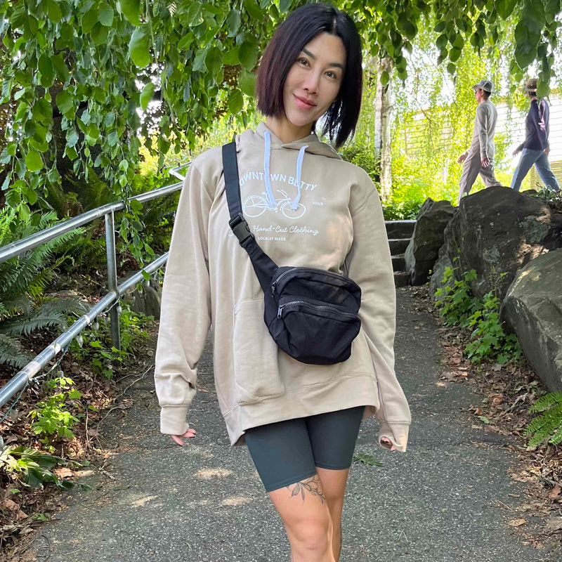 Person wearing a beige hoodie, bike shorts shorts, and white sneakers with a black bag, standing on a path with greenery.