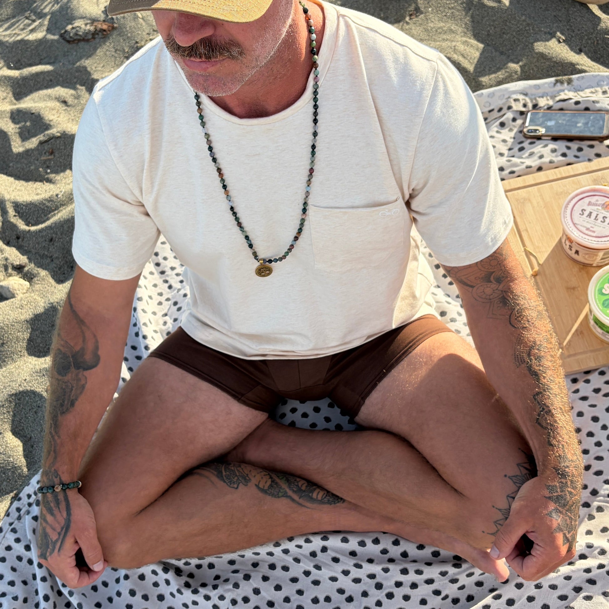 Man sitting on a blanket on a sandy beach with a picnic beside him.