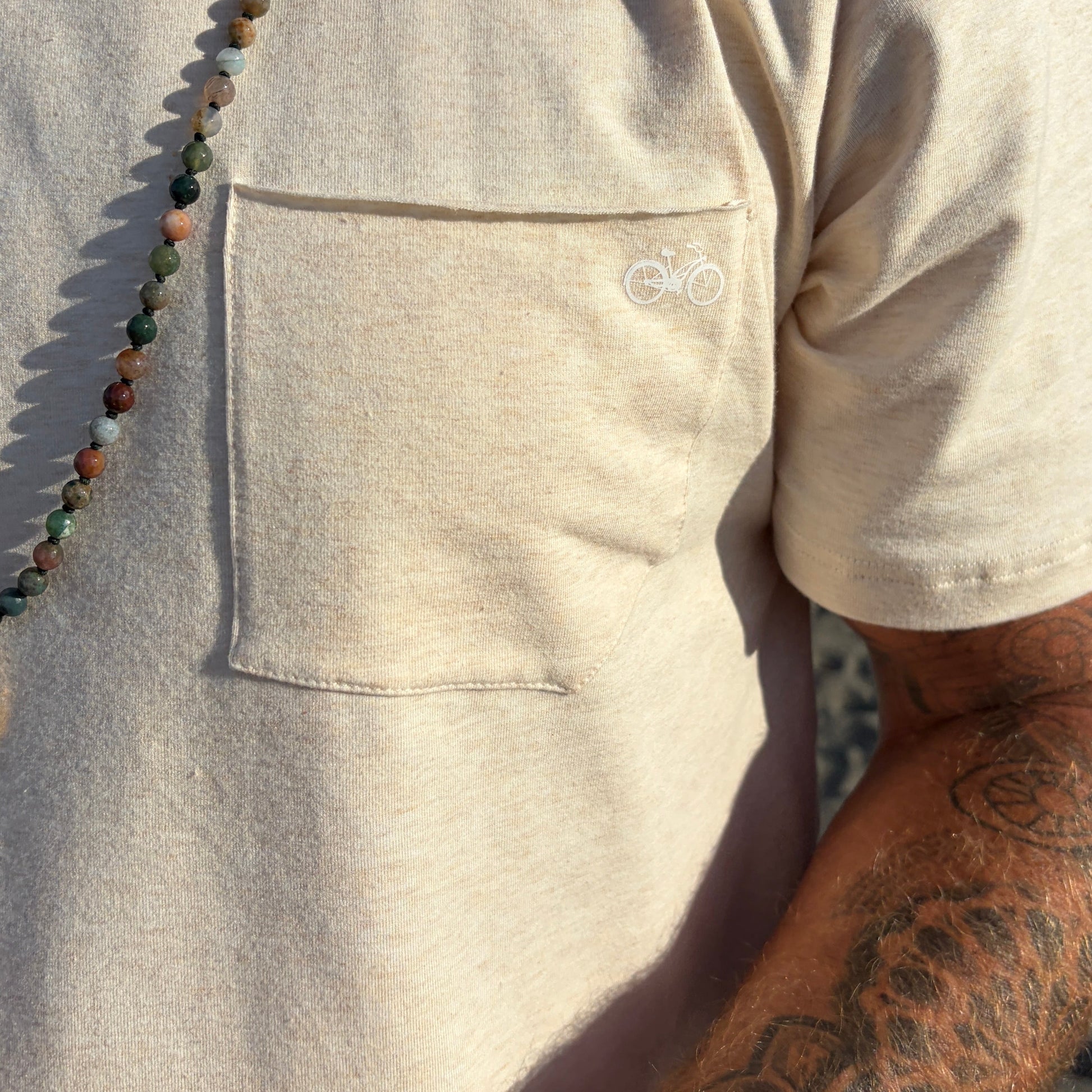 Person wearing a cream t-shirt with a pocket with a bicycle logo and a tattooed arm, standing outdoors.