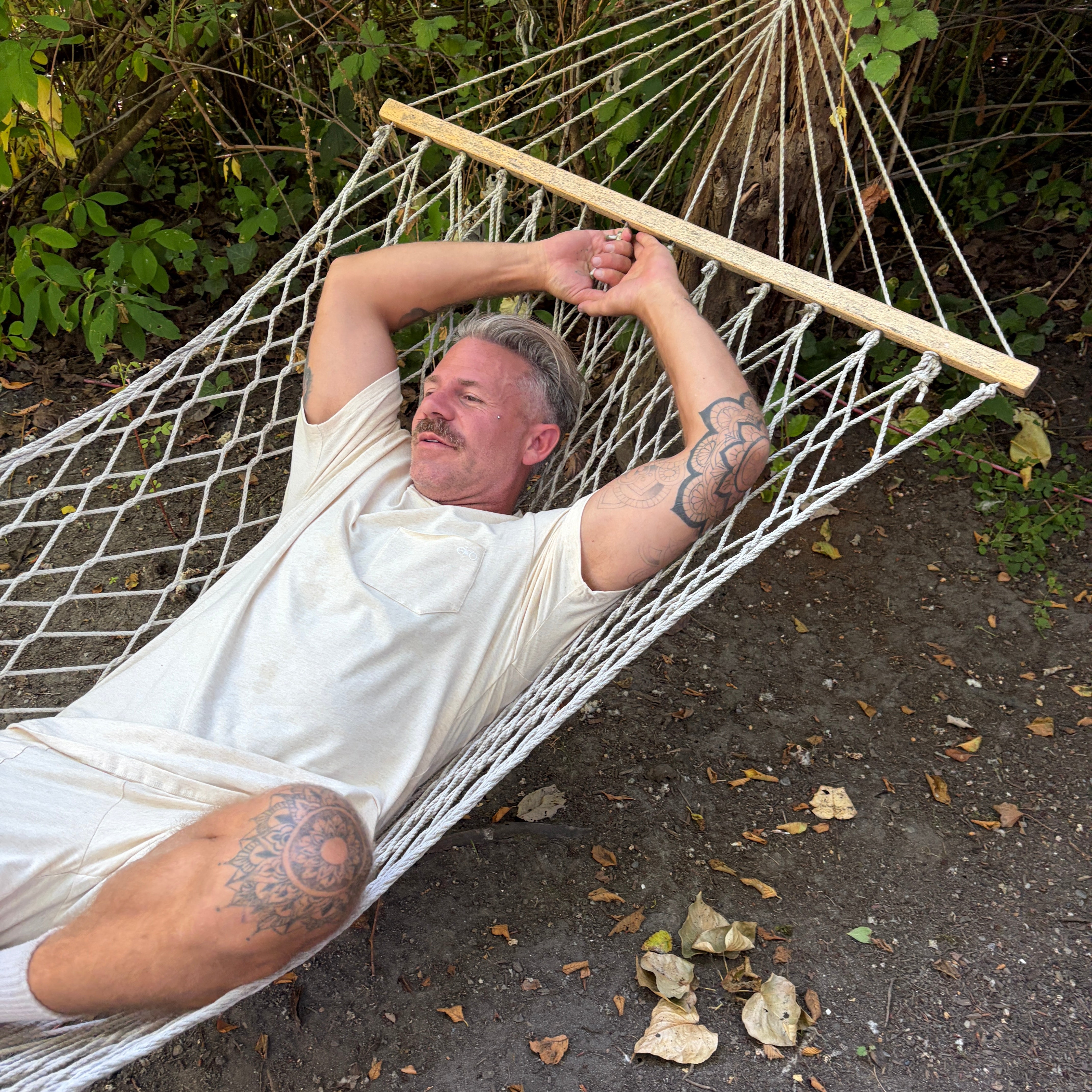 Man lying in a hammock outdoors wearing a cream outfit with trees and ground visible