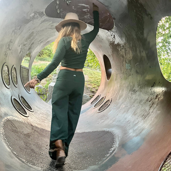 Person wearing a green outfit, standing inside a large metal tube structure with greenery in the background