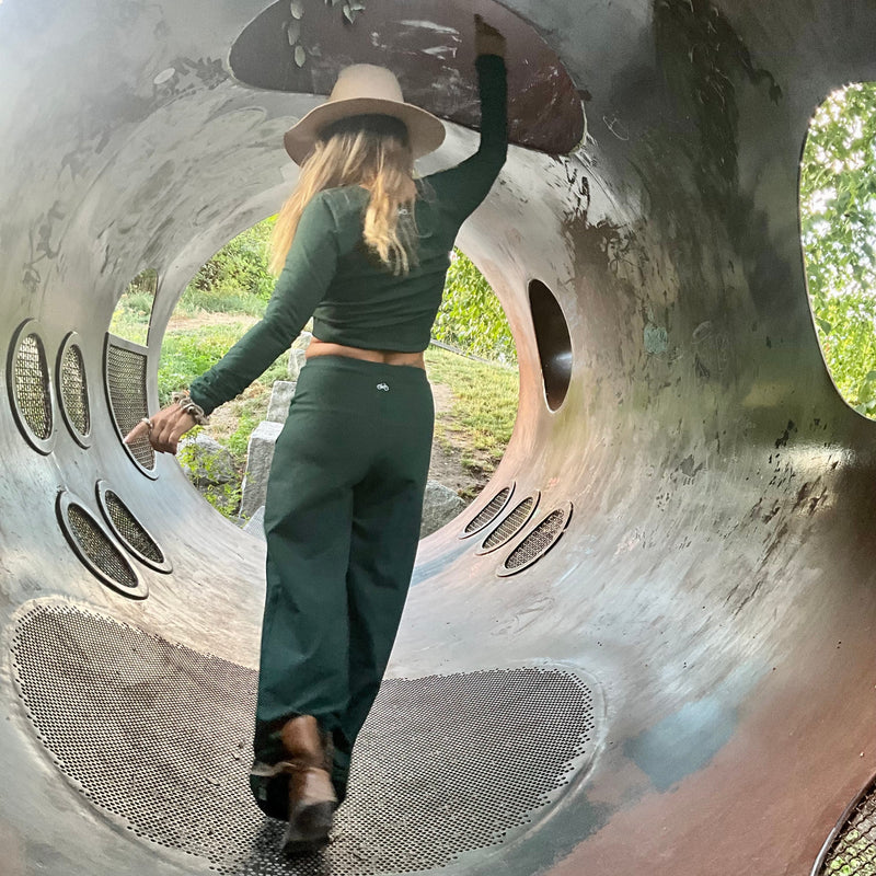 Person wearing a green outfit, standing inside a large metal tube structure with greenery in the background