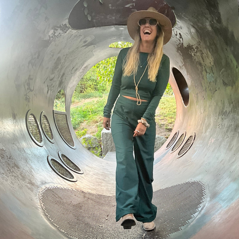Person laughing inside a large metal sculpture with a textured surface wearing green trousers & a fitted longsleeve crop top in emerald