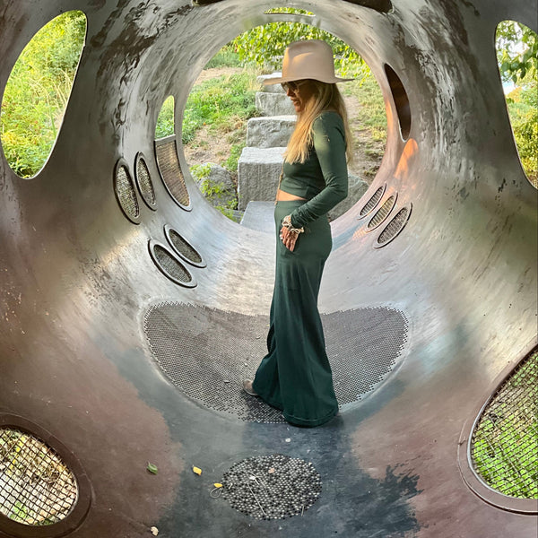 Woman in a longsleeve top & green pants standing inside a large, circular iron structure with natural light filtering through.