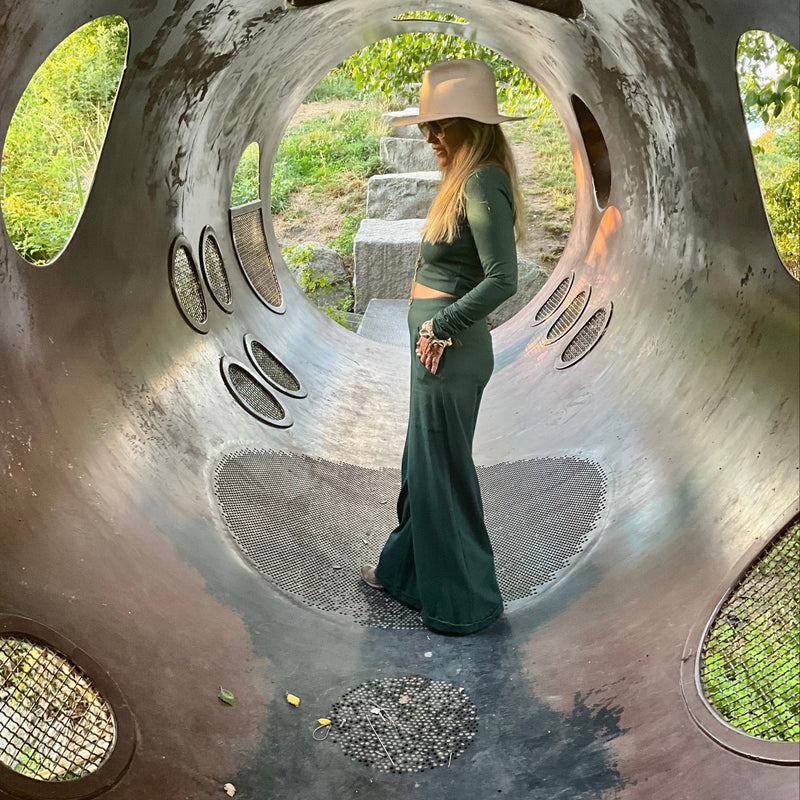Woman in a longsleeve top & green pants standing inside a large, circular iron structure with natural light filtering through.