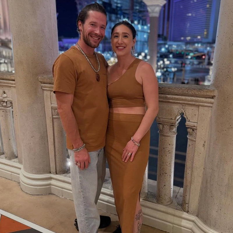 a couple posing for a photo in an elegant indoor setting wearing matching tobacco DowntownBetty SoftGoods outfits with a cityscape view.

