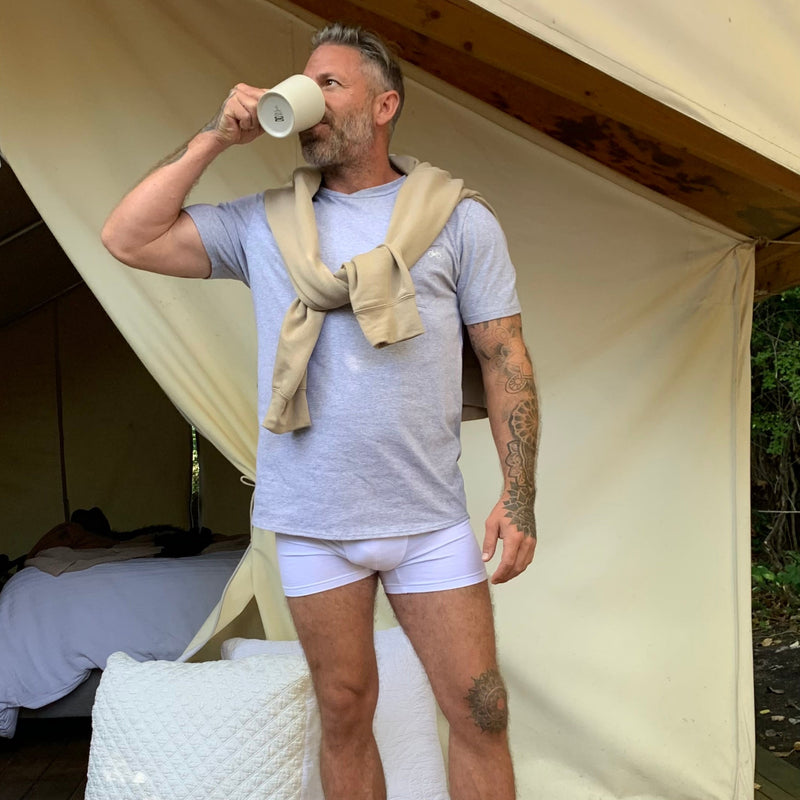 Man standing in front of a tent, wearing a light gray t-shirt, white underwear, and white socks, with a beigehoody over his shoulder.